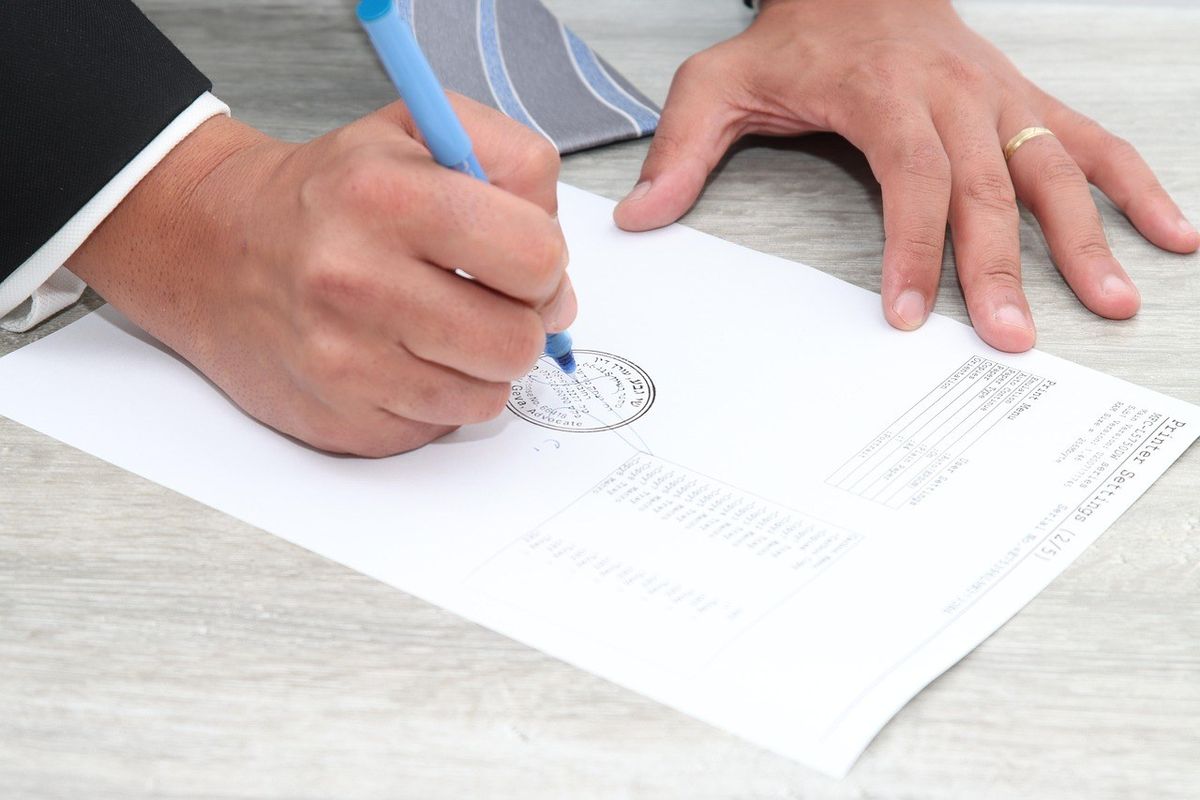 Main droite écrivant sur un document avec un stylo bleu, main gauche posée dessus, table en bois