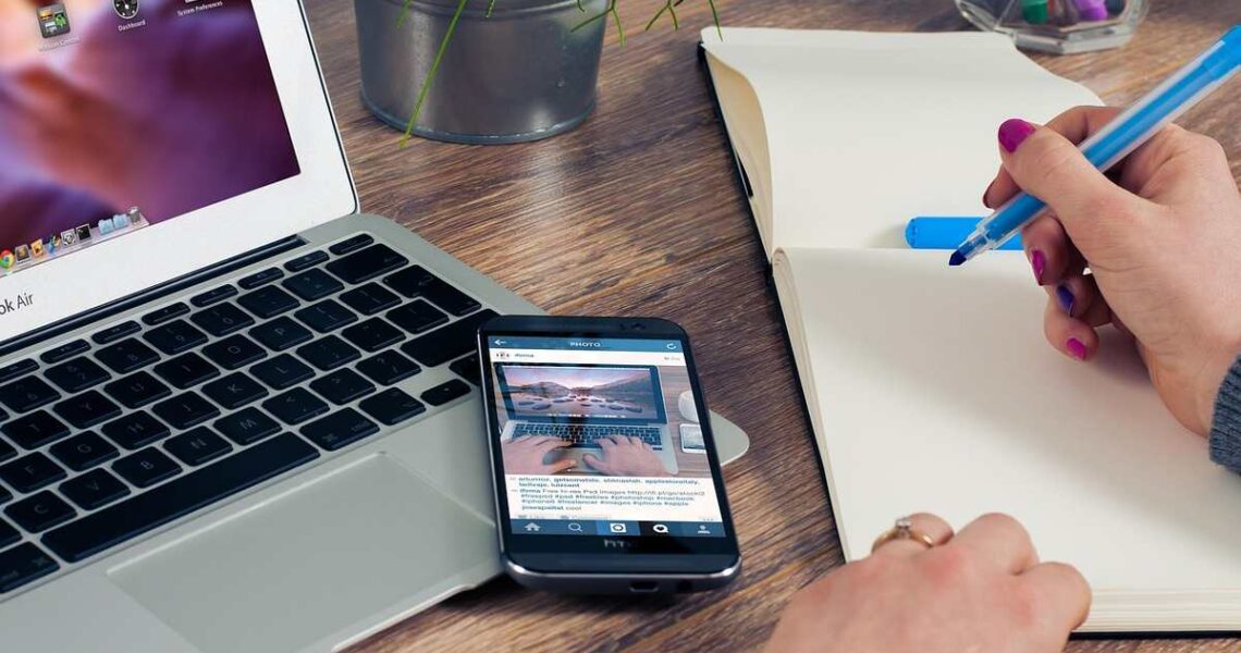 Une main écrit dans un cahier à côté d'un ordinateur portable et d'un smartphone