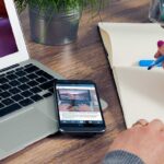 Une main écrit dans un cahier à côté d'un ordinateur portable et d'un smartphone