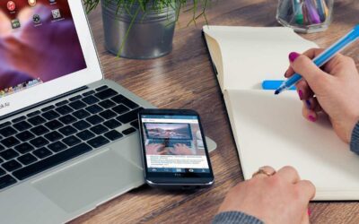 Une main écrit dans un cahier à côté d'un ordinateur portable et d'un smartphone